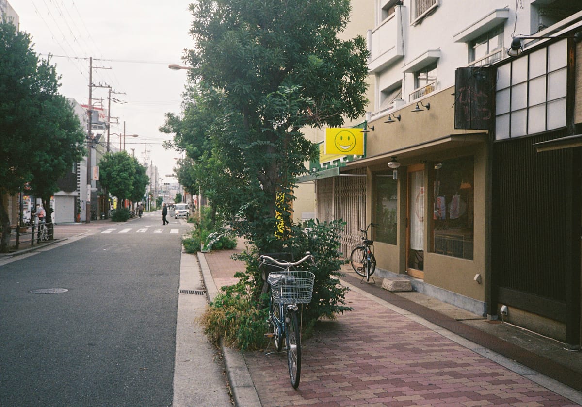 A street in Joto-ku, Osaka, with a restaurant and store ont he right, a few trees along the street, and a bicycle parked in front of a tree in the foreground. Photo was taken on film.